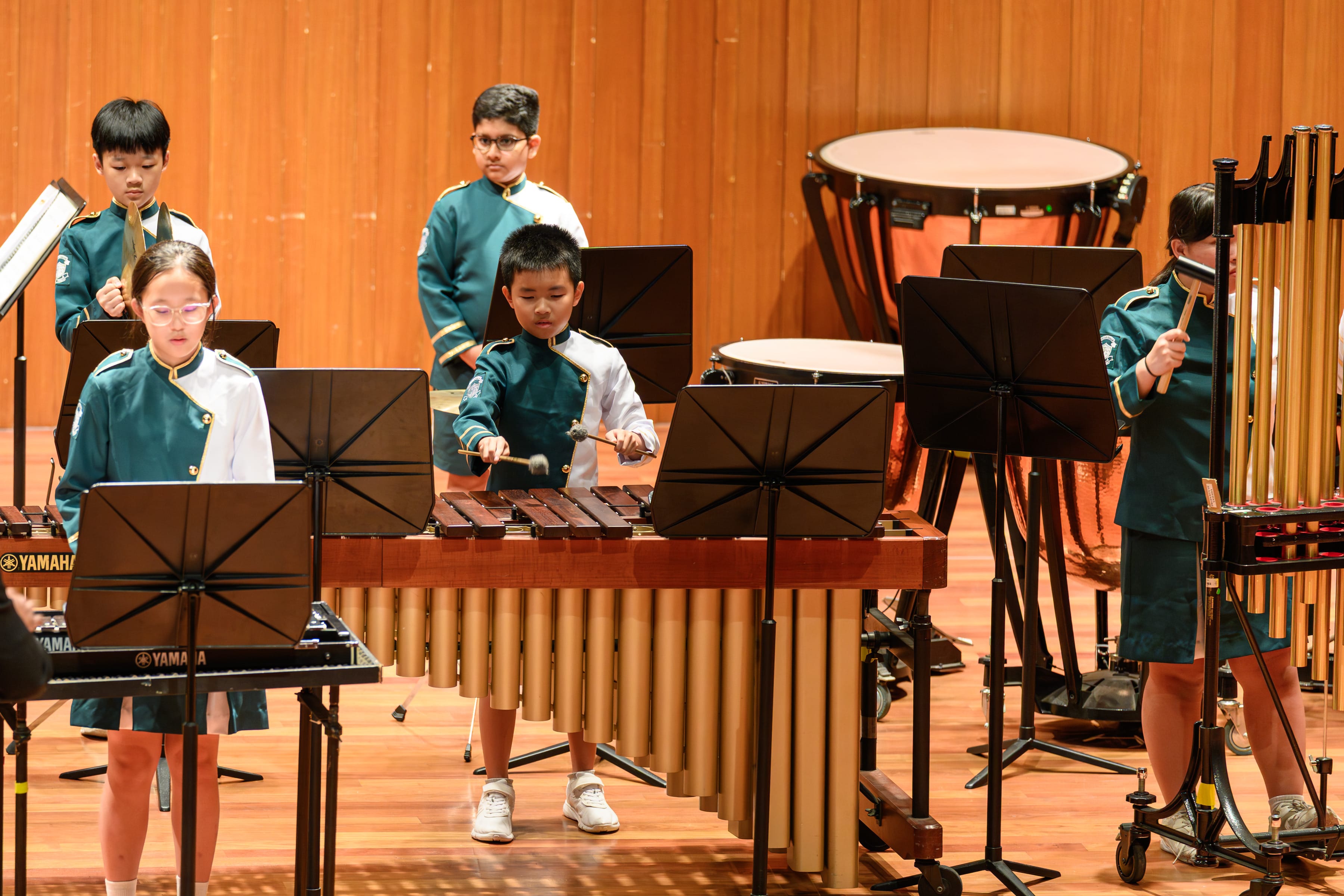 School band students in uniform performing a percussion ensemble on a concert stage, playing instruments including a marimba, xylophone, keyboard, and timpani, with music stands in front of them.